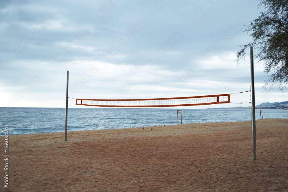 Landscape view on empty morning beach with volleyball grid stretched between two steel poles, dramatic cloudy sky and blue sea behind