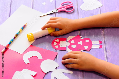 Small child made an angel doll of cardboard. Childrens hands on a wooden table. Craft supplies for a fun kids activity. Valentine's day crafts idea for kids in home or kindergarten