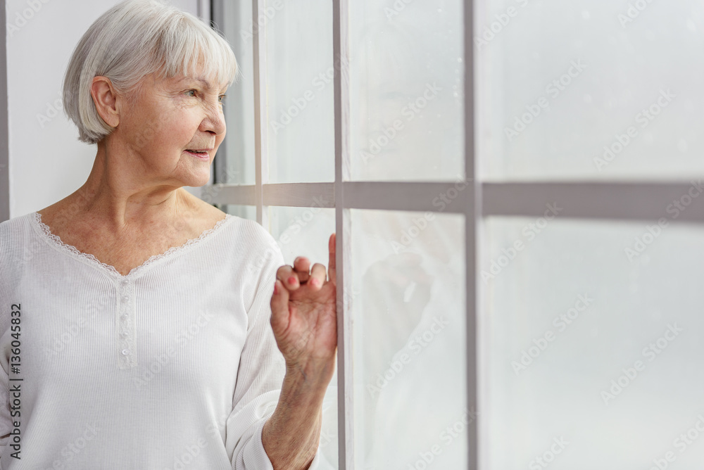 Cheerful pensioner watching at closed window in hospital Cheerful pensioner watching at closed window in hospital
