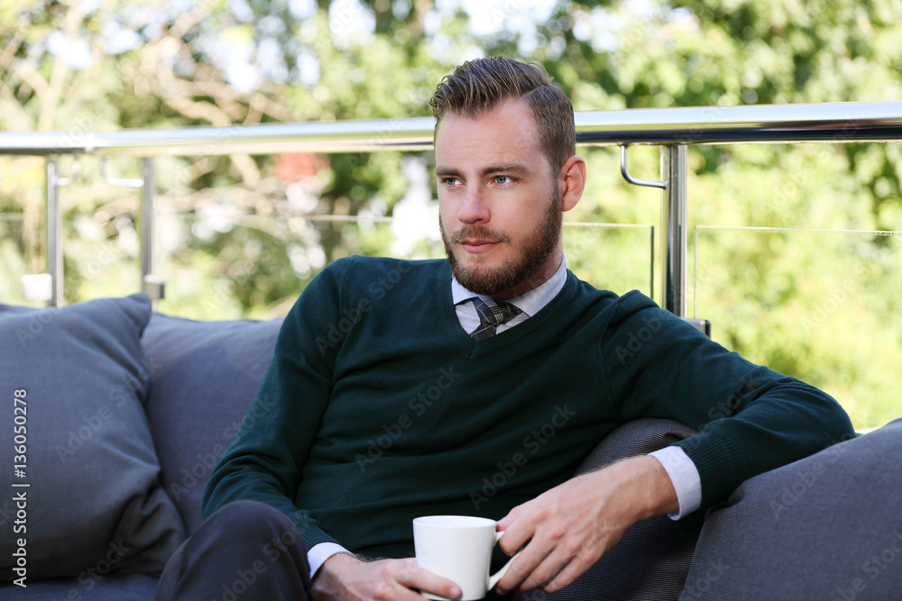 An attractive man in his 20s sitting down outdoors holding a mug looking away from camera, wearing a green pullover and shirt on a sunny summer day. An attractive man in his 20s sitting down outdoors holding a mug looking away from camera, wearing a green pullover and shirt on a sunny summer day.
