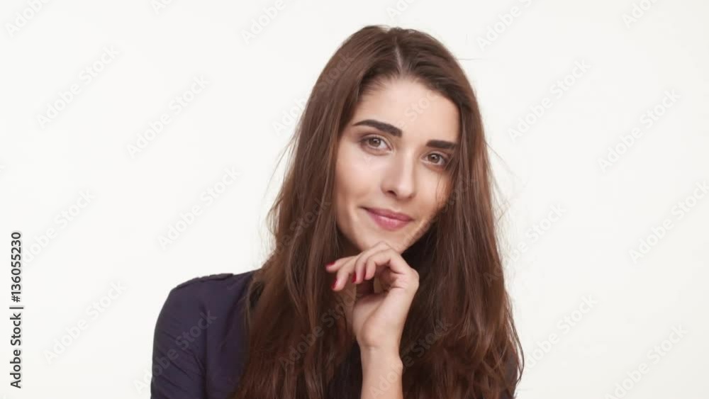 Shy coy beautiful Caucasian female with long brown hair standing on white background in dark blue shirt smiling touching hair in slowmotion Shy coy beautiful Caucasian female with long brown hair standing on white background in dark blue shirt smiling touching hair in slowmotion