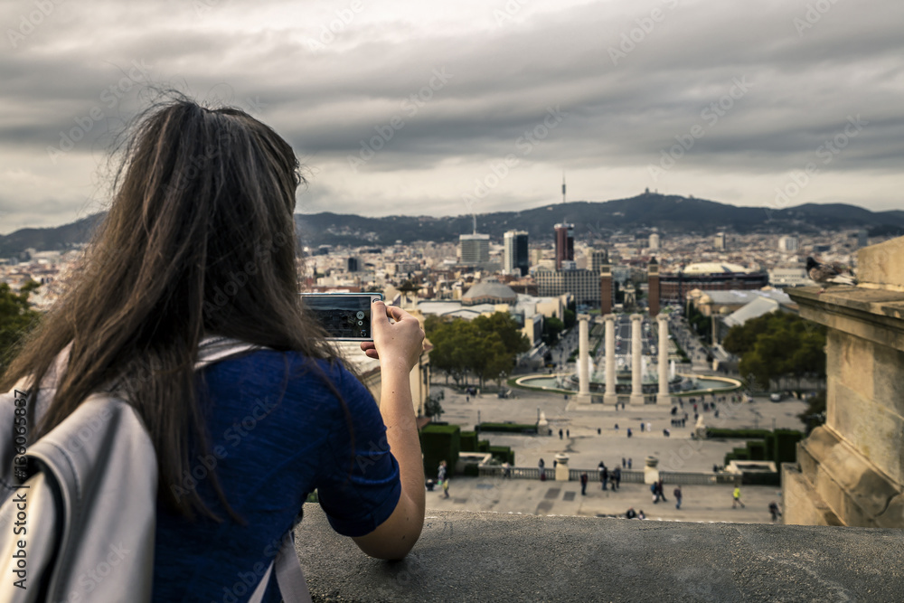 Young girl with long brown hair standing on view point and taking a picture of the city landscape using mobile phone. Backside view. Monjuic, Barcelona, Spain