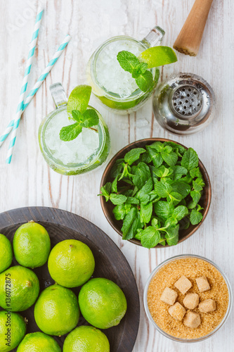 Two fresh mojitos cocktail in mason jar on wooden background. Mojitos with mint leaves, lime and ice. Drink making tools and ingredients for cocktail. View from above.