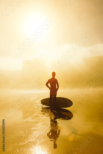 Surfer with his surfboard and looking for waves on a misty urban beach. Outdoor beach water sport and surf lifestyle.