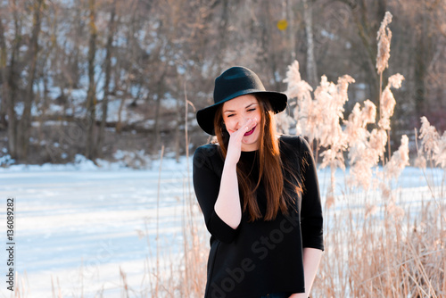 Winter portrait of a happy beautiful girl with brown hair in winter forest on the lake. Woman wearing a hat and bright clothes. Winter atmosphere, nature and freedom concept. 