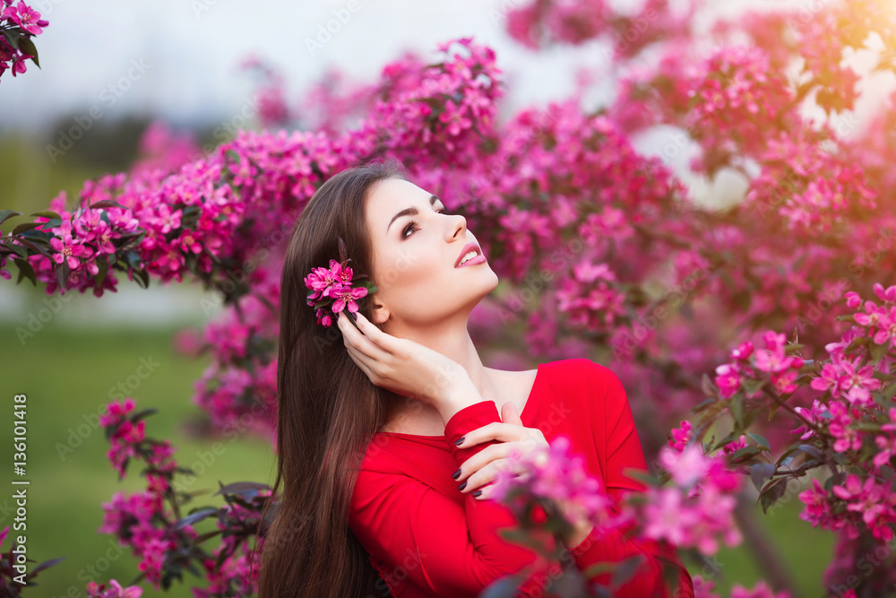 Spring touch. Happy beautiful young woman in red dress enjoy fresh pink flowers and sun light in blossom park at sunset.