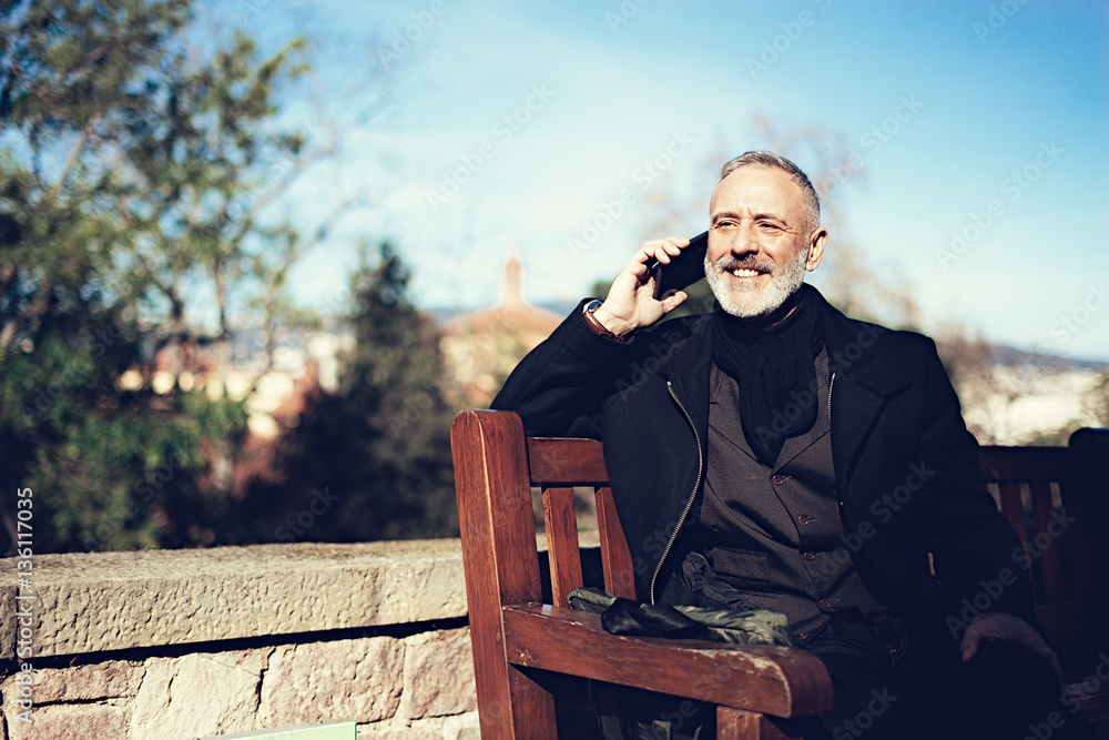 Fototapeta premium Smiling adult attractive businessman wearing a classic suit and talking at mobile phone while sitting in city park.Horizontal,blurred background.