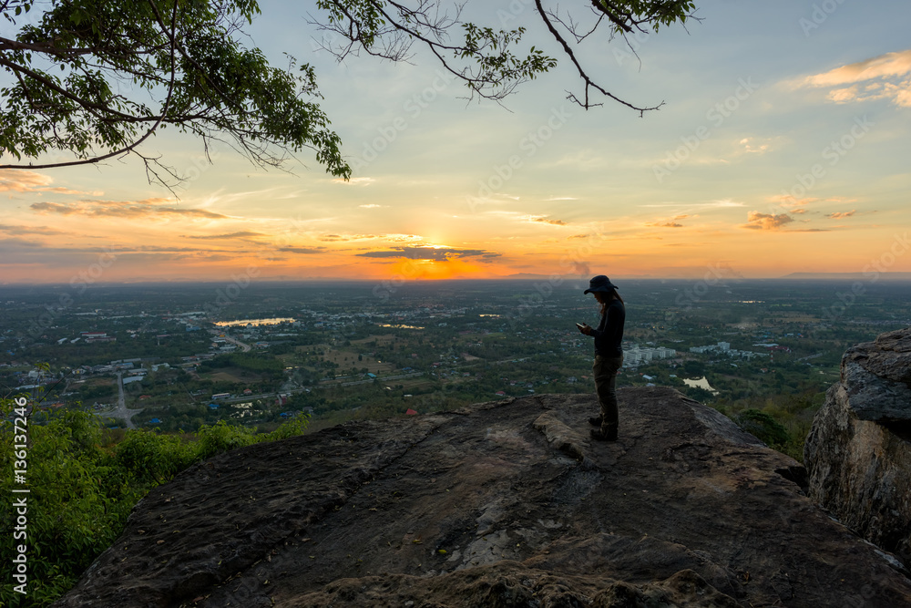 Man traveler is using digital phone on cliff with beautiful landscape sunset over cliff and city  - success concept