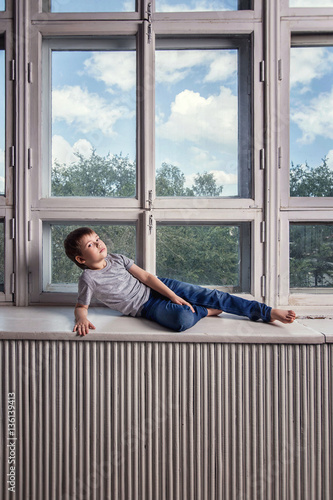 Little handsome boy in jeans and a grey shirt, barefoot, lying and posing on the windowsill of a huge white retro window