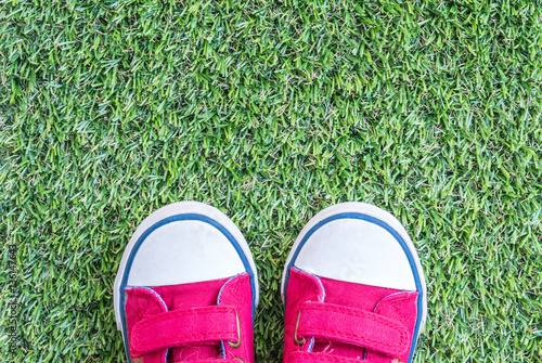 Closeup red fabric sneakers of kid on green artificial grass textured background in top view with copy space