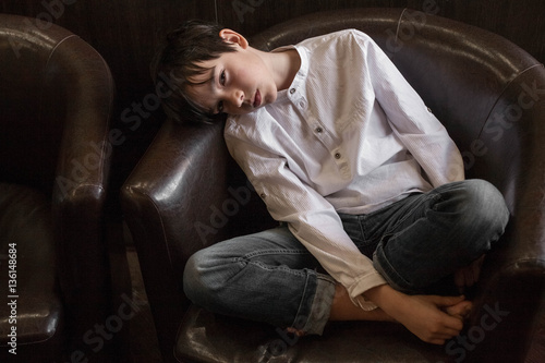 Boy in white shirt sitting in a chair. young boy imposingly placed in a leather couch. boy in shirt and jeans. Teenager relaxing in a cafe   © vetaka