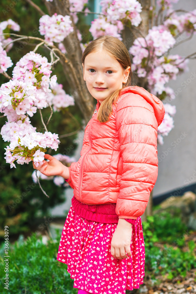 Spring portrait of a pretty little girl of 7 years old wearing coral jacket standing between branches of a blooming tree