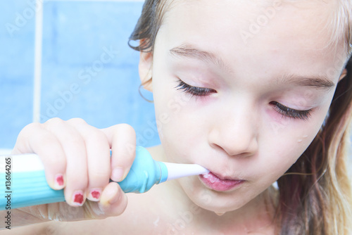 Close up of cute little girl in a bath and cleaning her teeth with electrical toothbrush against cool blue wall. Children hygiene.