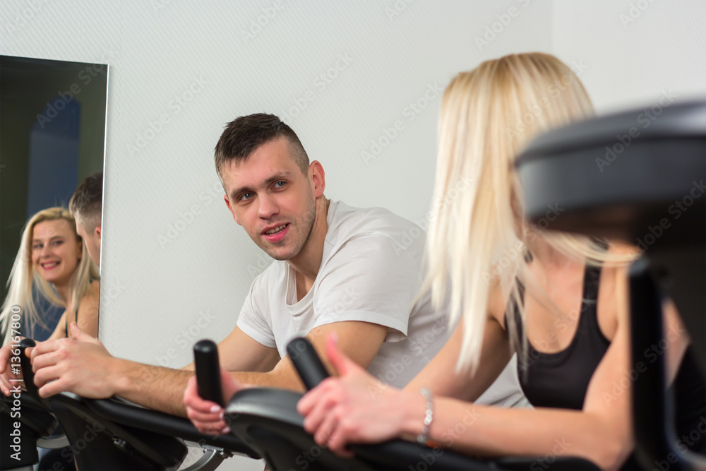 Obraz premium Young man and woman biking in the gym, exercising legs doing cardio workout cycling bikes