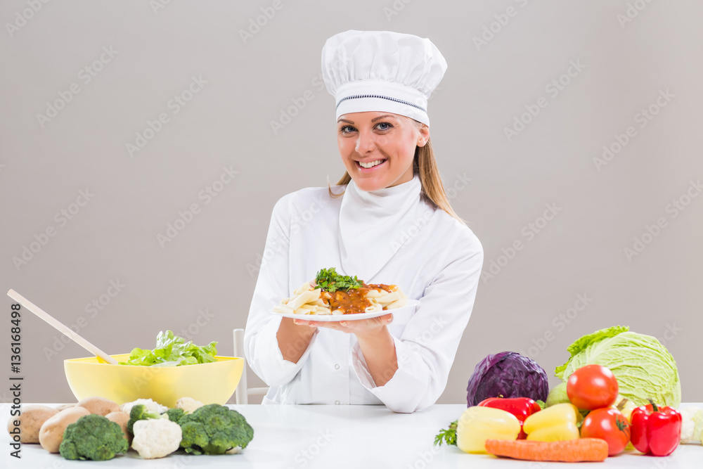 Cheerful female chef is sitting at the table with bunch of vegetable and showing prepared meal. Cheerful female chef is sitting at the table with bunch of vegetable and showing prepared meal.
