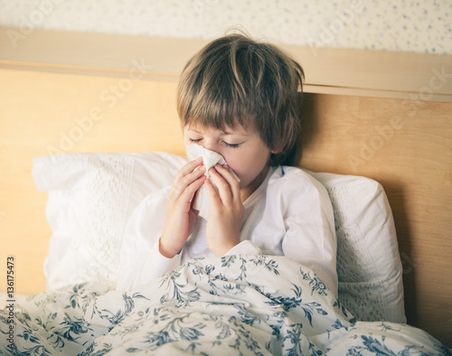 Unhappy ill kid lying in bed and blowing his nose 