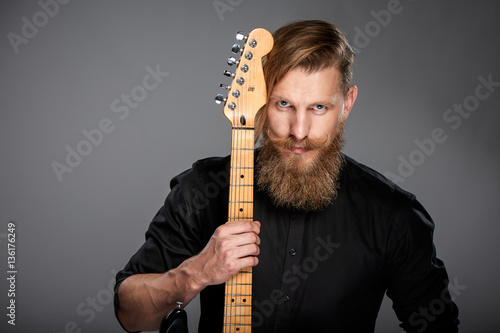 Closeup portrait of hipster man with beard and mustashes wearing black shirt holding guitar, over grey background photo