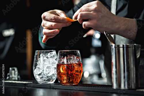 Closeup of bartender hands pouring alcoholic drink into a jigger to prepare a cocktail, with red bell pepper and peppercorn seeds in a serving glass.