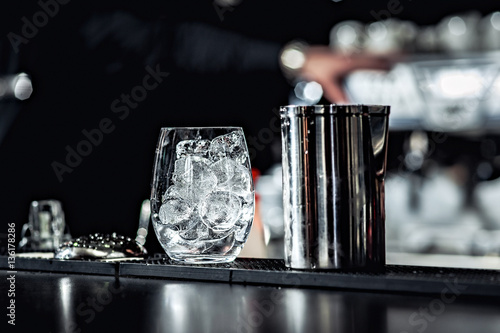 Closeup of bartender hands pouring alcoholic drink into a jigger to prepare a cocktail, with red bell pepper and peppercorn seeds in a serving glass.