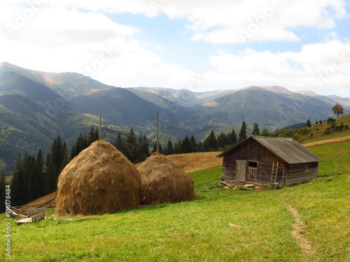 Obraz na plátně Old wooden hut and haystacks on  background of  beautiful mountain landscape and clouds