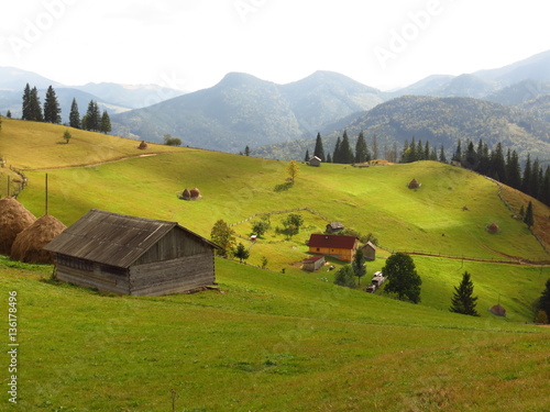 Fototapet Old wooden hut and haystacks on  background of  beautiful mountain landscape and clouds