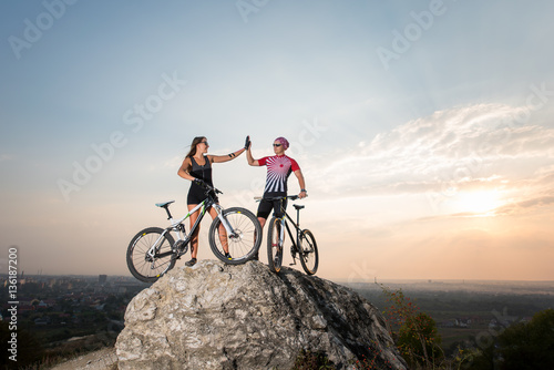 Man give high five for woman standing with the mountain bicycles on the rock under blue sky. Below is a small city in the distance
