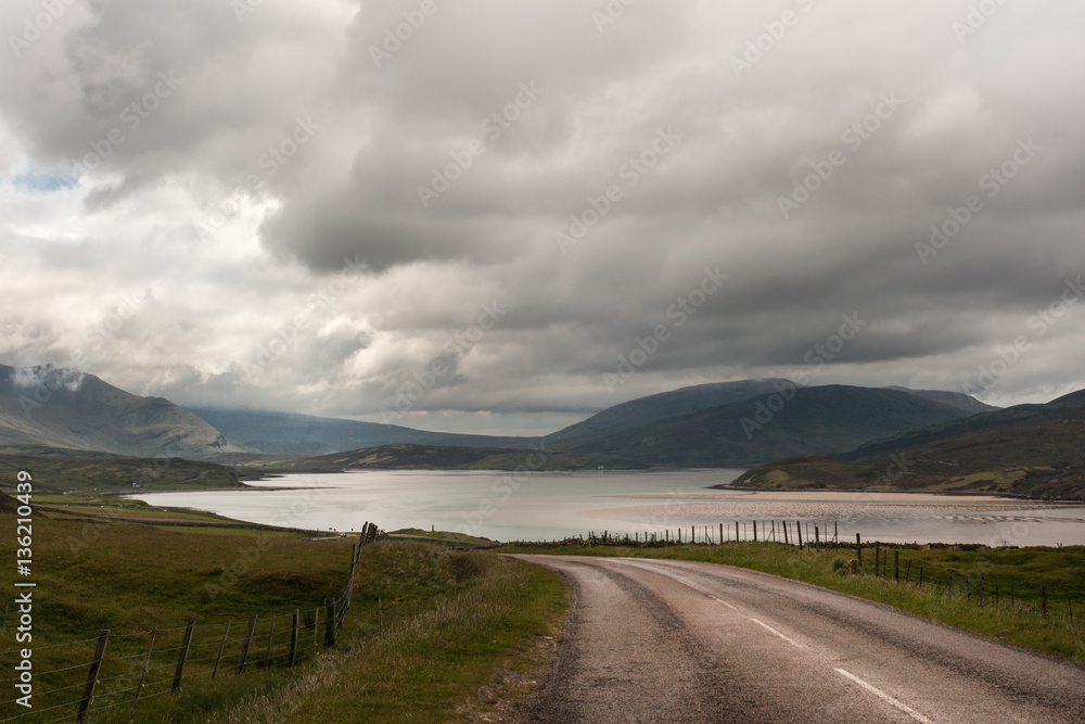 Northwest Coast, Scotland - June 6, 2012: The rural road called highway descends on Kyle of Durness, part of Atlantic Ocean. Heavy dark and white clouds. Mountains around. Low tide shows sand banks.