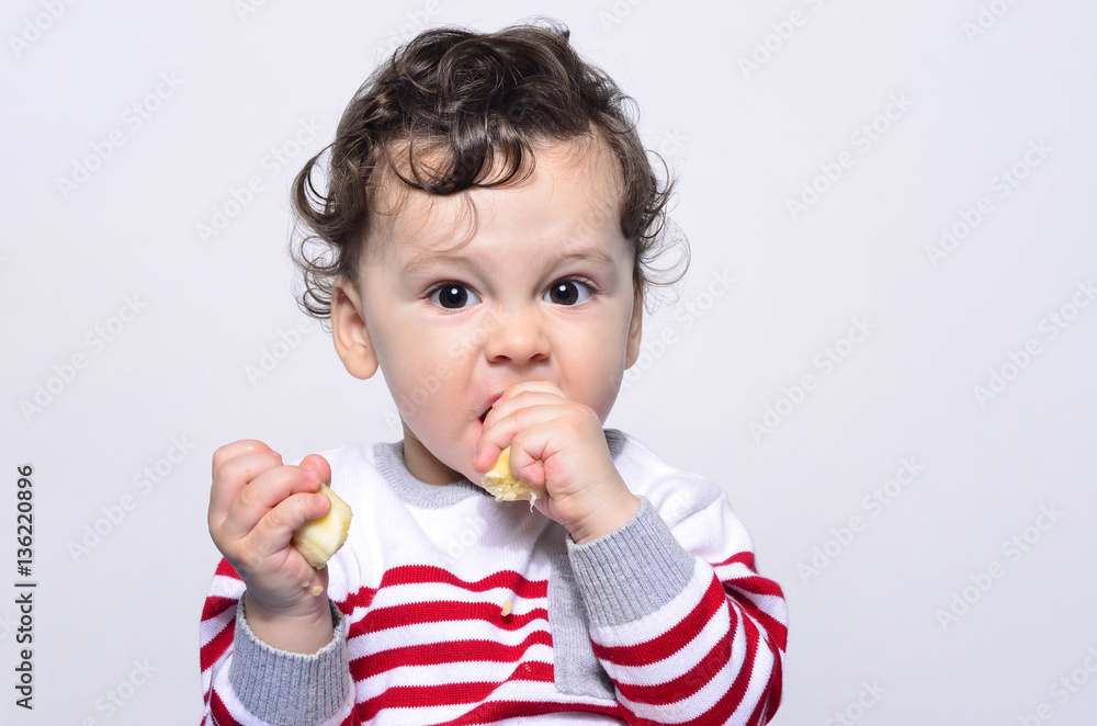 Portrait of a cute baby eating a banana.One year old kid eating fruits by himself. Adorable curly hair boy being hungry. Portrait of a cute baby eating a banana.One year old kid eating fruits by himself. Adorable curly hair boy being hungry.