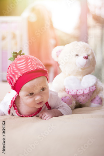 Close up of a beautiful baby girl lying on the bed. Infant lying on a bed. Family morning at home. Happy baby wearing strawberry shirt and pants. Newborn kid with toy bear.