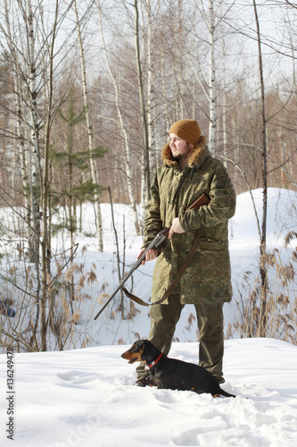 Hunter man with young black smooth-haired dachshund and old 16 caliber side-by-side double-barreled shotgun dressed in camouflage clothing in winter forest