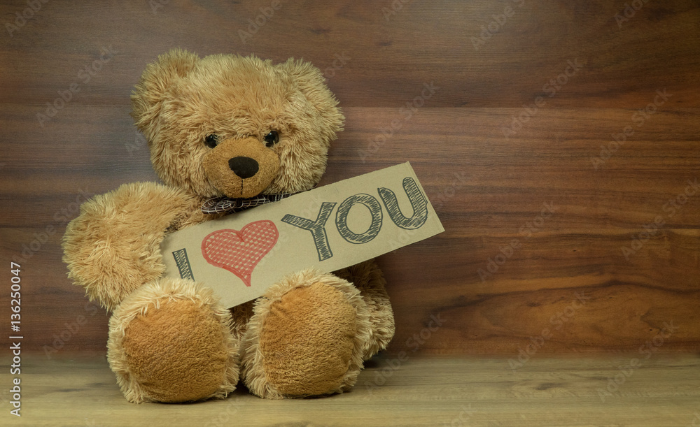 Obraz premium Cute fluffy teddy bear holding a cardboard I Love You sign, shot on a beautiful wooden surface with enough room for your message