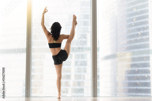 Woman practicing yoga, standing in Utthita Hasta Padangustasana exercise, Variation of Extended Hand to Big Toe pose, working out, wearing black tank top, shorts, full length, near window, rear view