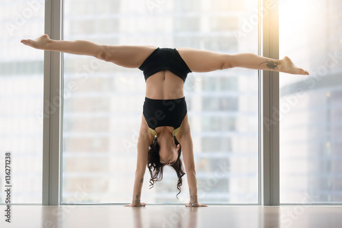 Young modern dancer attractive woman practicing, doing handstand exercise, dance pose, working out, wearing sportswear, black tank top, shorts, indoor full length, near floor window with city view