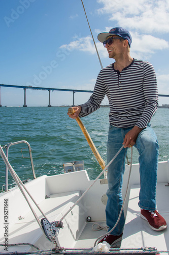 A man in a striped sweater, jeans, a blue cap and red sneakers controls sailing boat. Boy holds the tiller of a sailing boat in the hands. The bridge on background. Adventure. Sailor