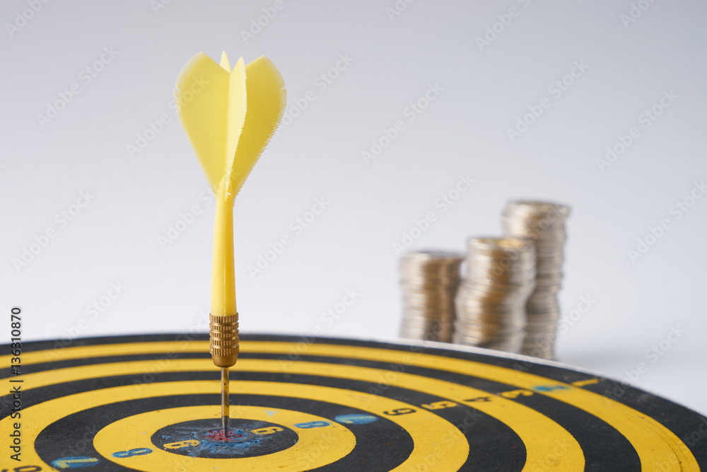 Yellow dart hit in the center of a target with stack of coins behind on white background with copy space. A idea about money / currency investment that must decide or think carefully Yellow dart hit in the center of a target with stack of coins behind on white background with copy space. A idea about money / currency investment that must decide or think carefully