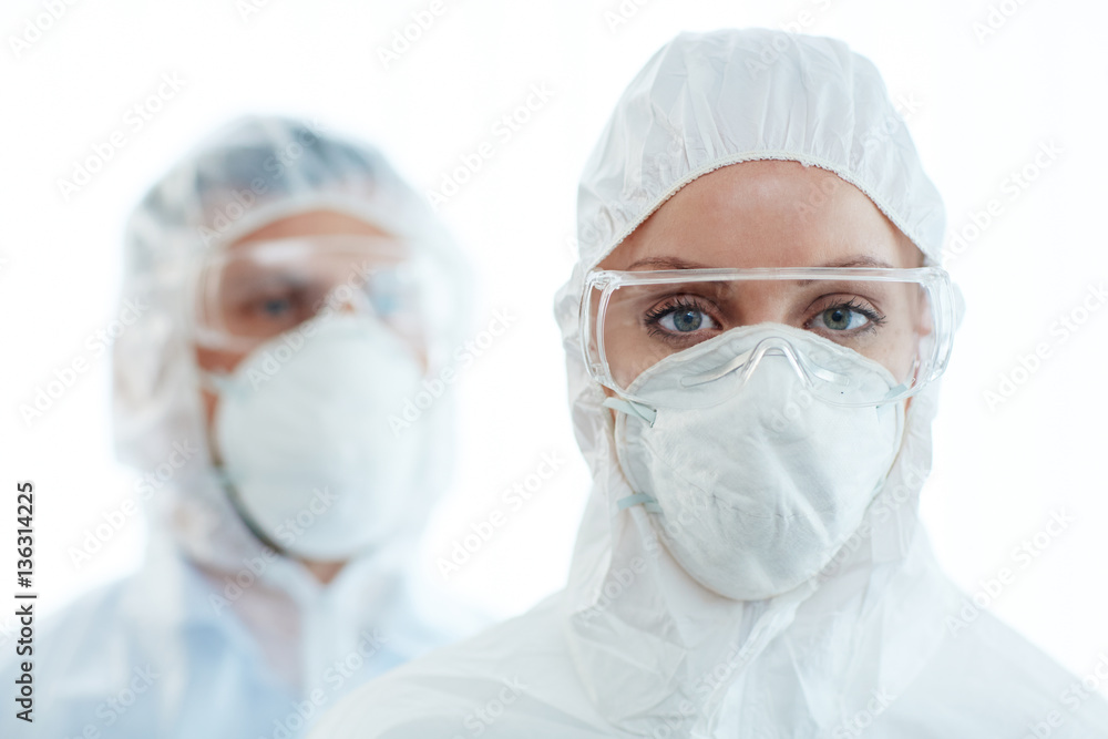 Young female in coveralls, eyeglasses and respirator looking at camera with her colleague on background
