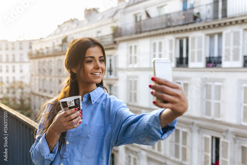 Young woman standing on balcony taking smart phone selfie photo
