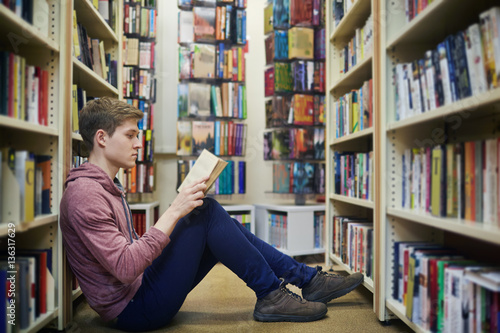 Serious guy sitting on the floor between bookshelves and reading in library