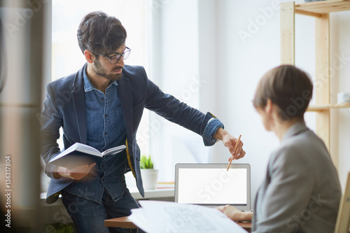 Young stylish manager helping female colleague with work issues pointing to blank laptop screen and explaining plans in modern office