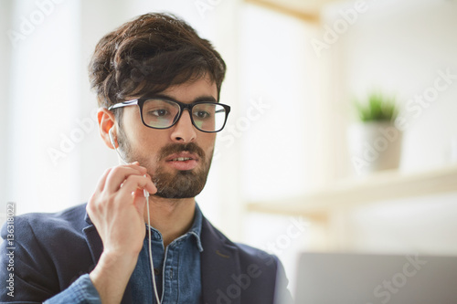 Portrait of young creative man wearing glasses focused while holding videochat interview applying for job position using hands free mic and laptop at modern workplace