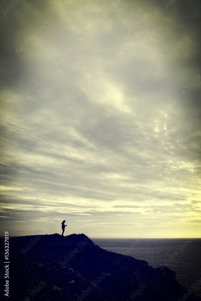 Vintage style photograph of a young woman standing on a cliff at sunset looking at her mobile phone