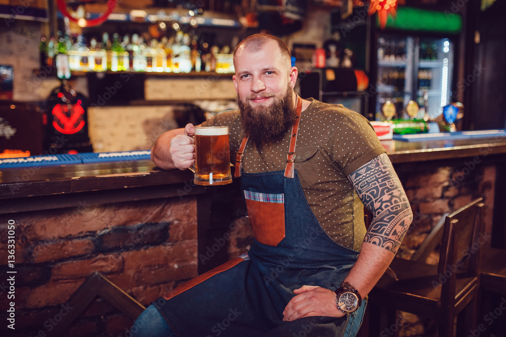 Bearded barman with tattoos wearing an apron sitting near the bar and holding a glass of beer