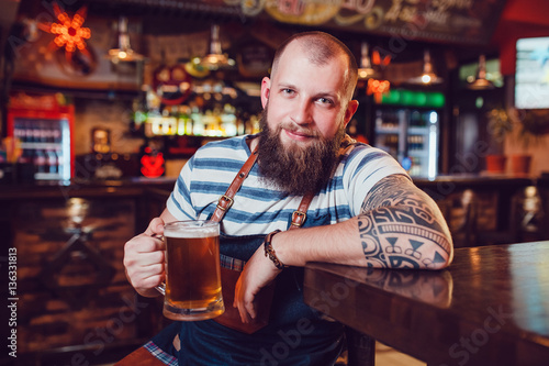 Bearded barman with tattoos wearing an apron sitting at the bar and holding a glass of beer.