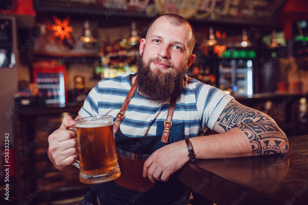 Bearded barman with tattoos wearing an apron sitting at the bar and holding a glass of beer.