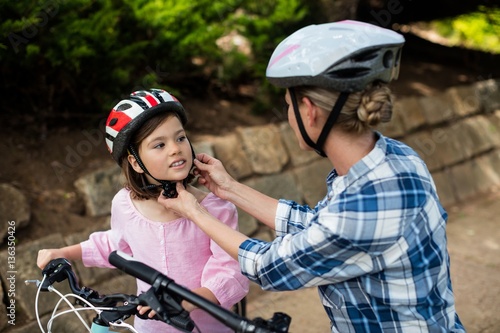 Mother assisting daughter in wearing bicycle helmet in park