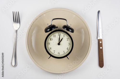 On the plate is an alarm clock, lying next to a knife and fork, white background, top view