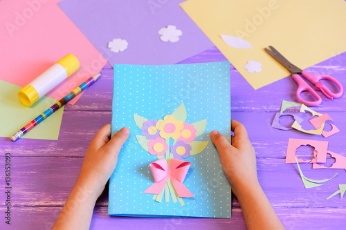 Small child made a greeting card with flowers for mom. Child holds a card in his hands. Tools and materials for children's art creativity on table. Mother's day or March 8 greeting card DIY idea