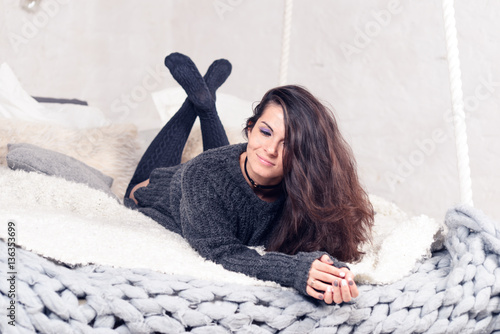 Beautiful sexy tan brunette woman posing in bedroom, wearing cozy grey sweater and knitted socks. Rest, sleeping, comfort concept. Close up of young woman lying in bed at home. Scandinavian home. 