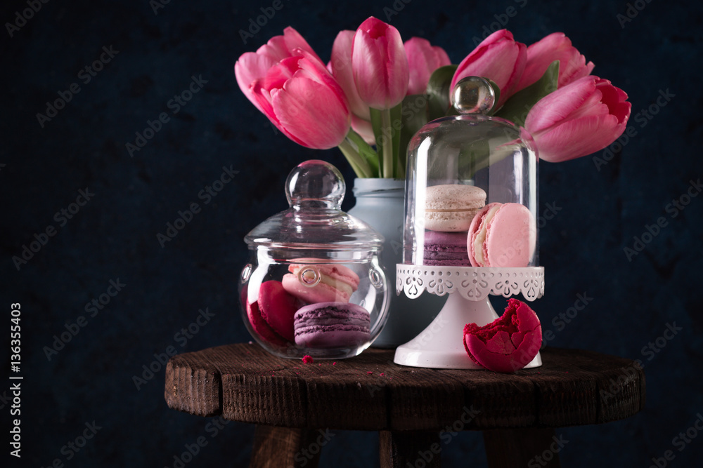Colorful tasty macaroons in glass jars and pink tulips on dark blue wall background Colorful tasty macaroons in glass jars and pink tulips on dark blue wall background