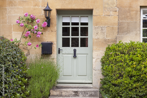 Light green wooden doors in an old traditional English lime stone cottage surrounded by climbing pink roses, lavender, on summer day © Yols
